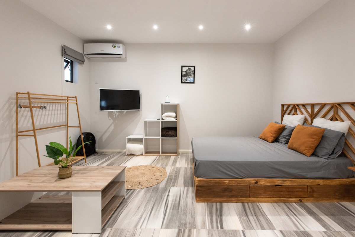 A modern studio space features a spacious bed with neutral linens and accent pillows. A small coffee table sits in front of the bed, accompanied by a decorative plant. Shelving units provide storage, while a wall-mounted television and air conditioning unit enhance comfort.