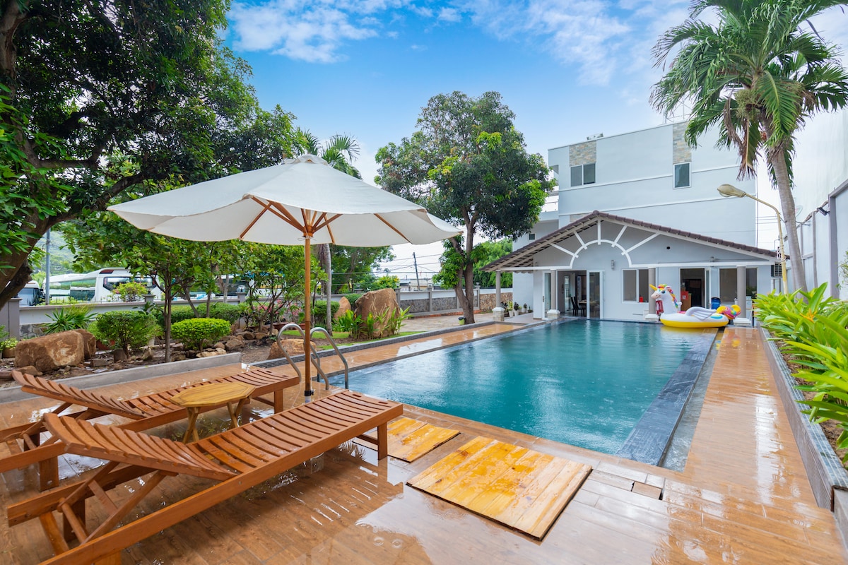 An outdoor space features a large swimming pool surrounded by wooden decking. Two lounge chairs are positioned under a large umbrella. Lush greenery and trees provide shade, while a spacious house is visible in the background.