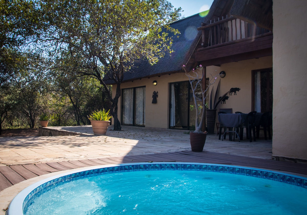 The image captures a serene outdoor space featuring a swimming pool surrounded by a wooden deck. The home is characterized by large glass doors, providing views of the lush greenery. A dining area with a table is also visible, situated under the shade of trees.