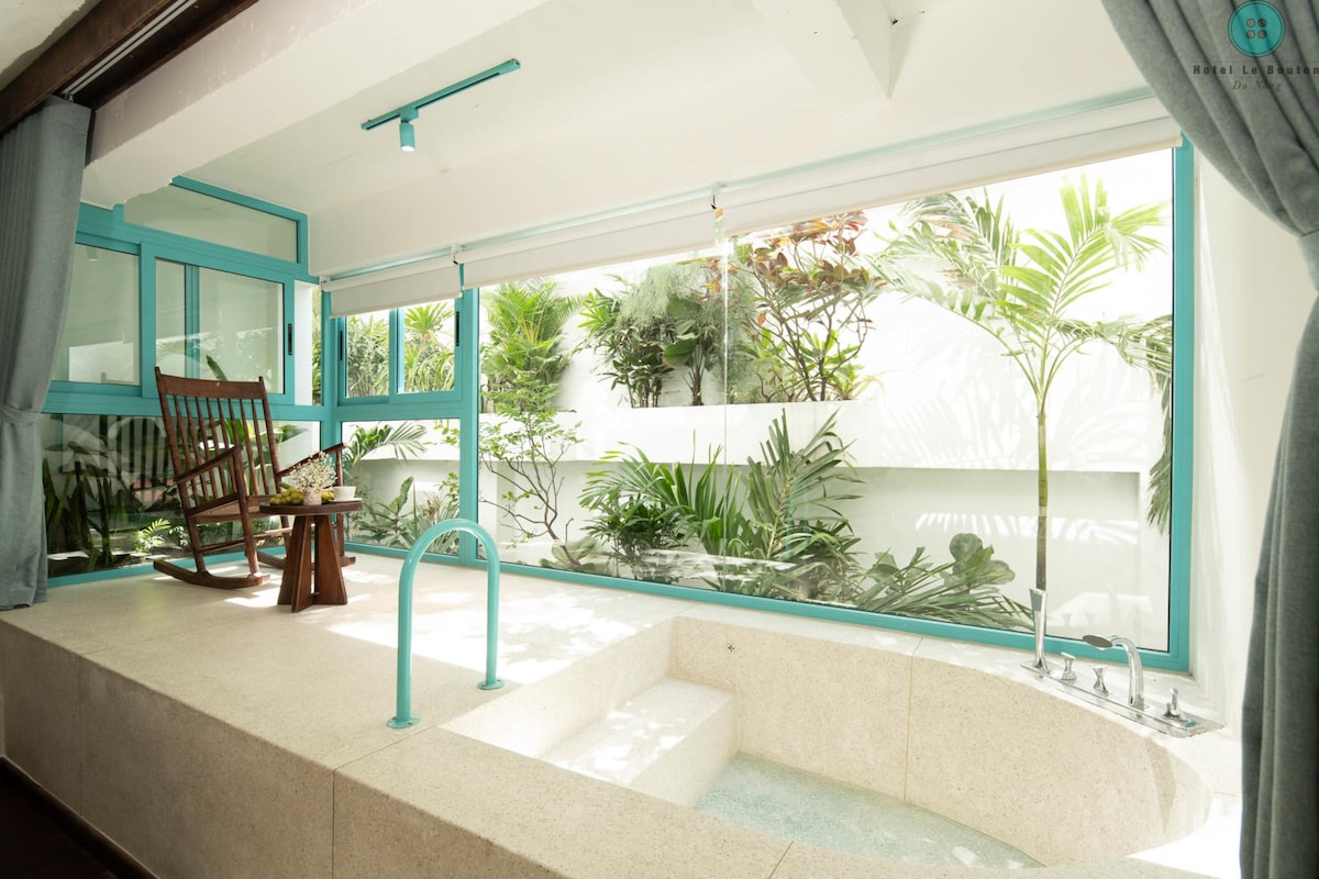 A bright and airy relaxation area features large windows that frame views of a lush garden. A spacious bathtub is positioned beside a comfortable wooden rocking chair. Soft curtains blend with the light decor, creating a serene space that invites tranquility.