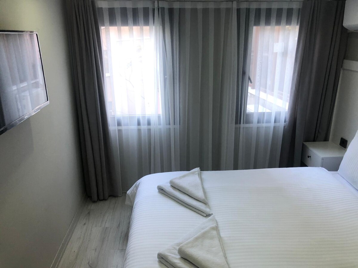 A well-lit sleeping area features a spacious bed draped with crisp linens. Two large windows adorned with sheer curtains allow natural light to fill the room, while a flat-screen television is mounted on the wall across from the bed.