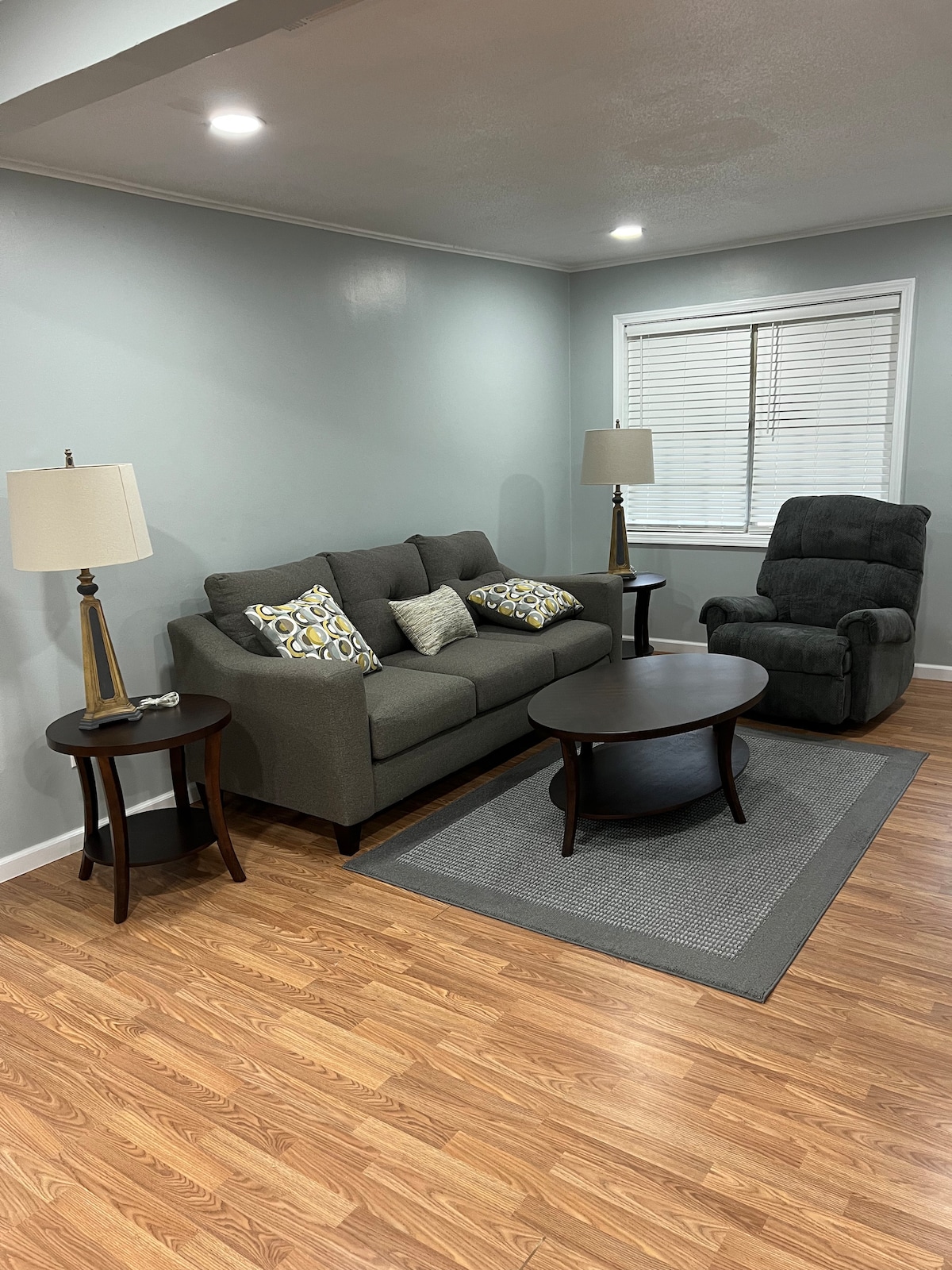 A cozy living area is furnished with a gray sofa adorned with decorative pillows. A round coffee table is positioned in the center, complemented by two side tables topped with lamps. A reclining chair is situated nearby, and natural light streams in through a window with blinds.