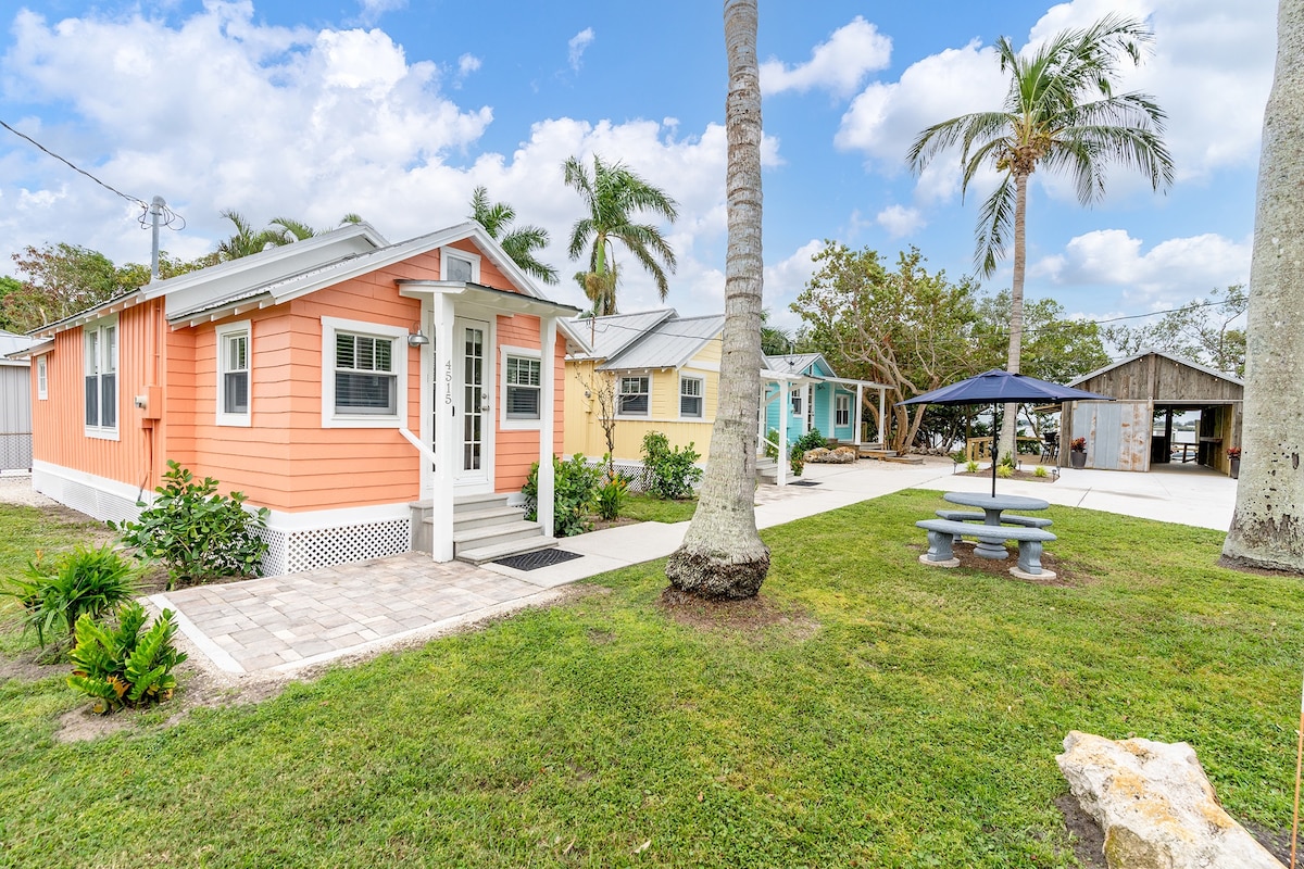 A charming waterfront cottage, featuring a peach-colored exterior, is surrounded by palm trees and green grass. A patio with a picnic table is visible in the foreground, and the property includes additional cottages in pastel colors. The area reflects a clean and inviting outdoor ambiance.