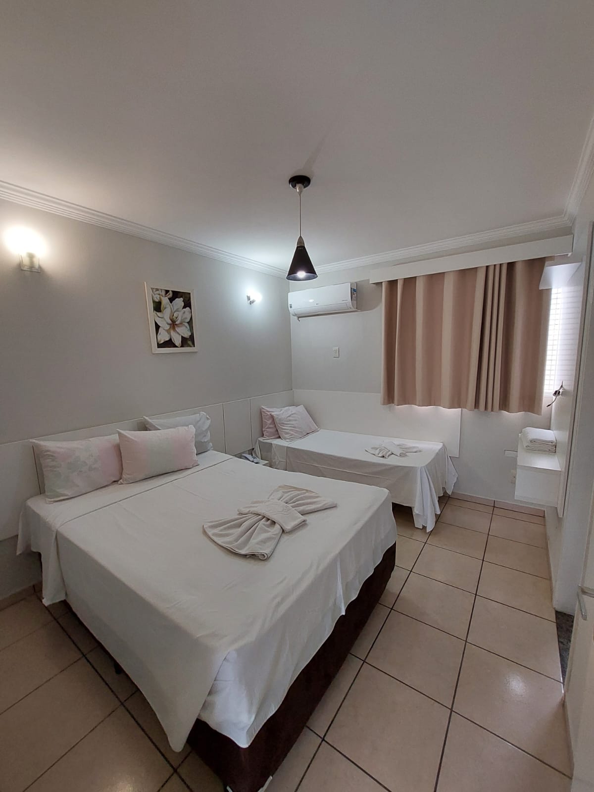 A well-lit room features a spacious bed with light bedding and decorative pillows, complemented by a cozy daybed nearby. Soft curtains filter natural light from a window, while a floral artwork adorns the wall, creating a serene environment.