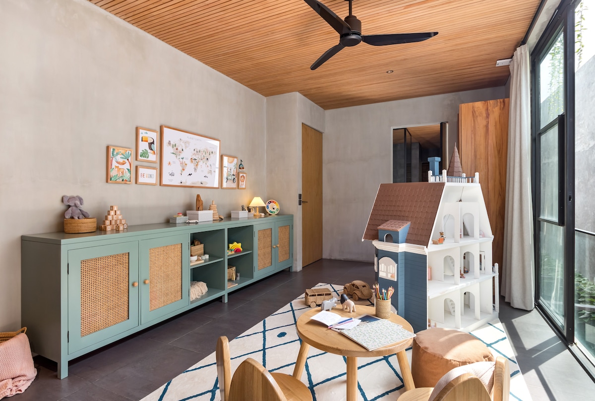 A playful kids' room features light grey walls and warm wood ceiling accents. A soft area rug with geometric patterns lies on the floor, surrounded by a wooden table and chairs. Colorful toys and a dollhouse occupy the space, enhancing a fun and inviting environment.