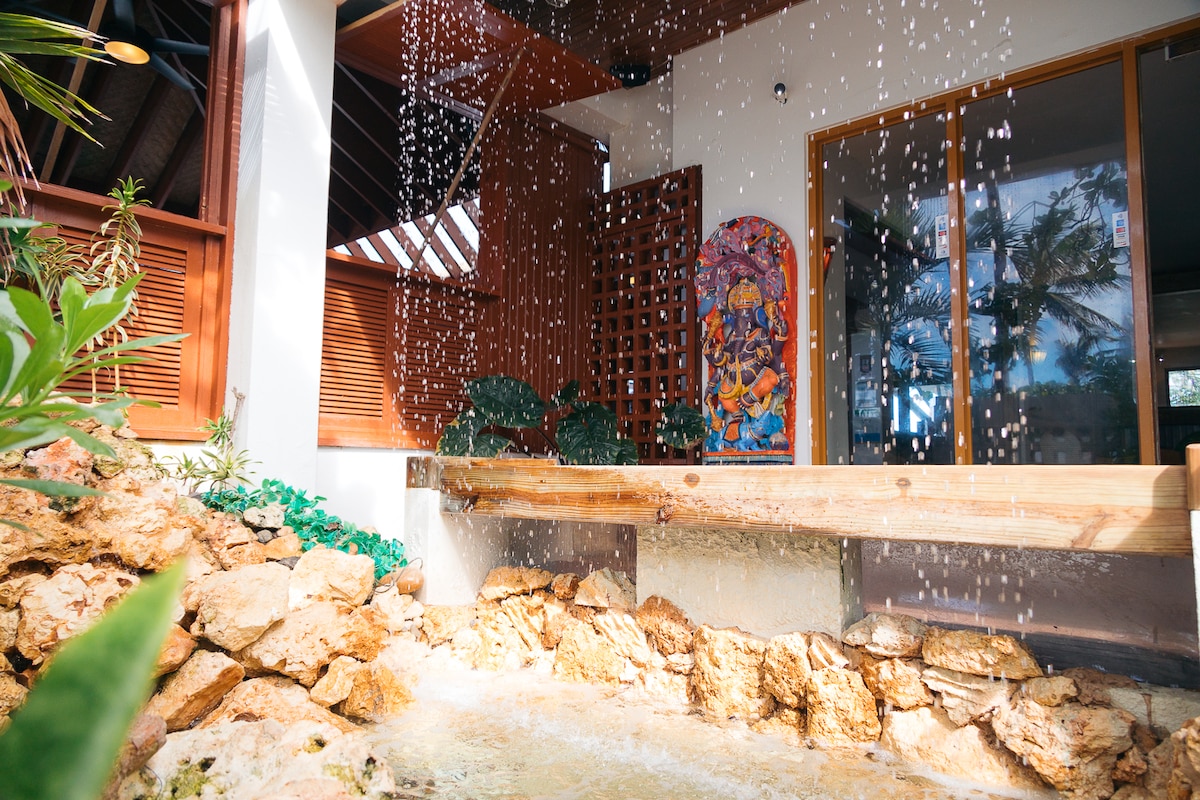 A natural water feature flows gently over textured stone, creating a serene ambiance. The background showcases large windows allowing light to enter, and a vibrant piece of wall art adds a splash of color among lush greenery.