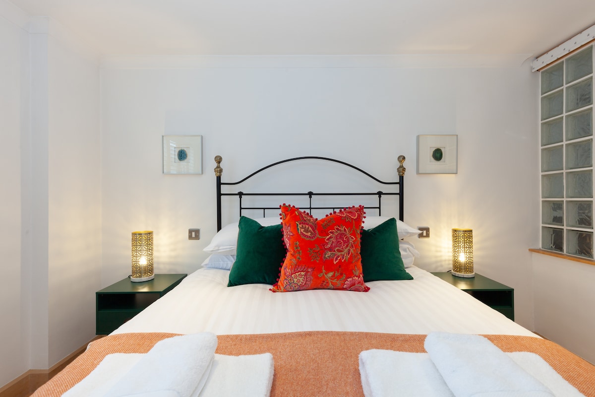 A spacious bed is centered against a simple white wall, embellished with decorative pillows in vibrant hues. Flanking the bed are two nightstands, each topped with stylish lamps. A soft orange throw adds warmth, while rolled towels are neatly placed at the foot.