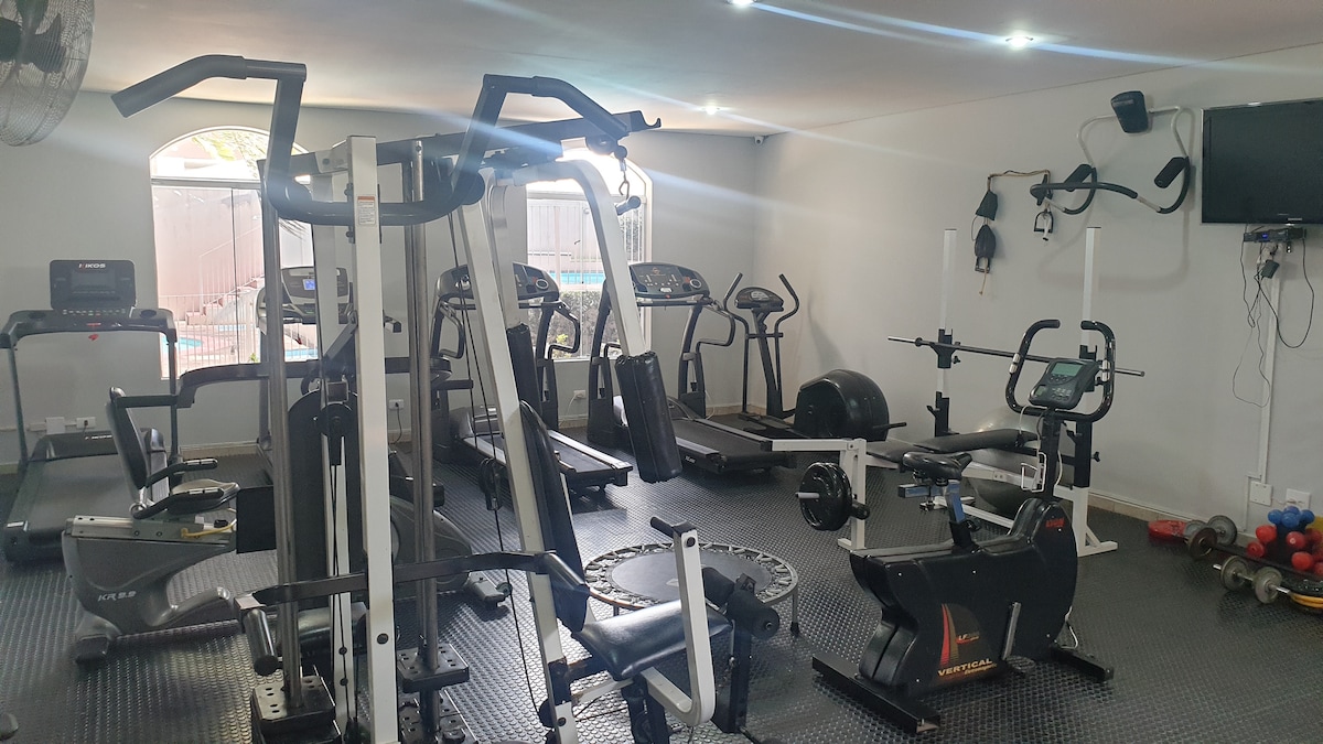 A well-equipped fitness area is presented with various exercise machines, including treadmills, elliptical trainers, and stationary bikes. Free weights and a stability ball are positioned in the corner, and large windows allow natural light to brighten the space.