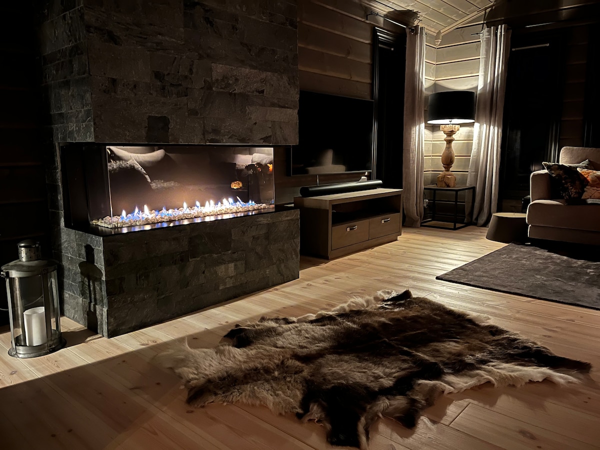 A modern interior features a sleek fireplace with a glass front, showcasing a gentle flame. Natural wood flooring is complemented by a textured, dark area rug. Soft lighting from a stylish lamp enhances the cozy ambiance, while a comfortable seating area is visible to the side.