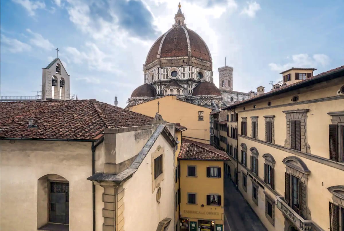 The image captures a view of the iconic Florence Cathedral, known as the Duomo, with its distinctive dome dominating the skyline. Surrounding buildings feature a mix of architectural styles, with terracotta roofs and elegant facades. Clear skies provide a bright backdrop to this historic scene.