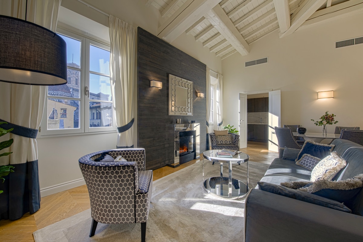 A spacious living room features a comfortable seating arrangement with patterned chairs and a plush sofa. Natural light flows in through large windows, offering a view of the iconic Duomo. A modern coffee table sits at the center, while a decorative fireplace adds to the inviting ambiance.