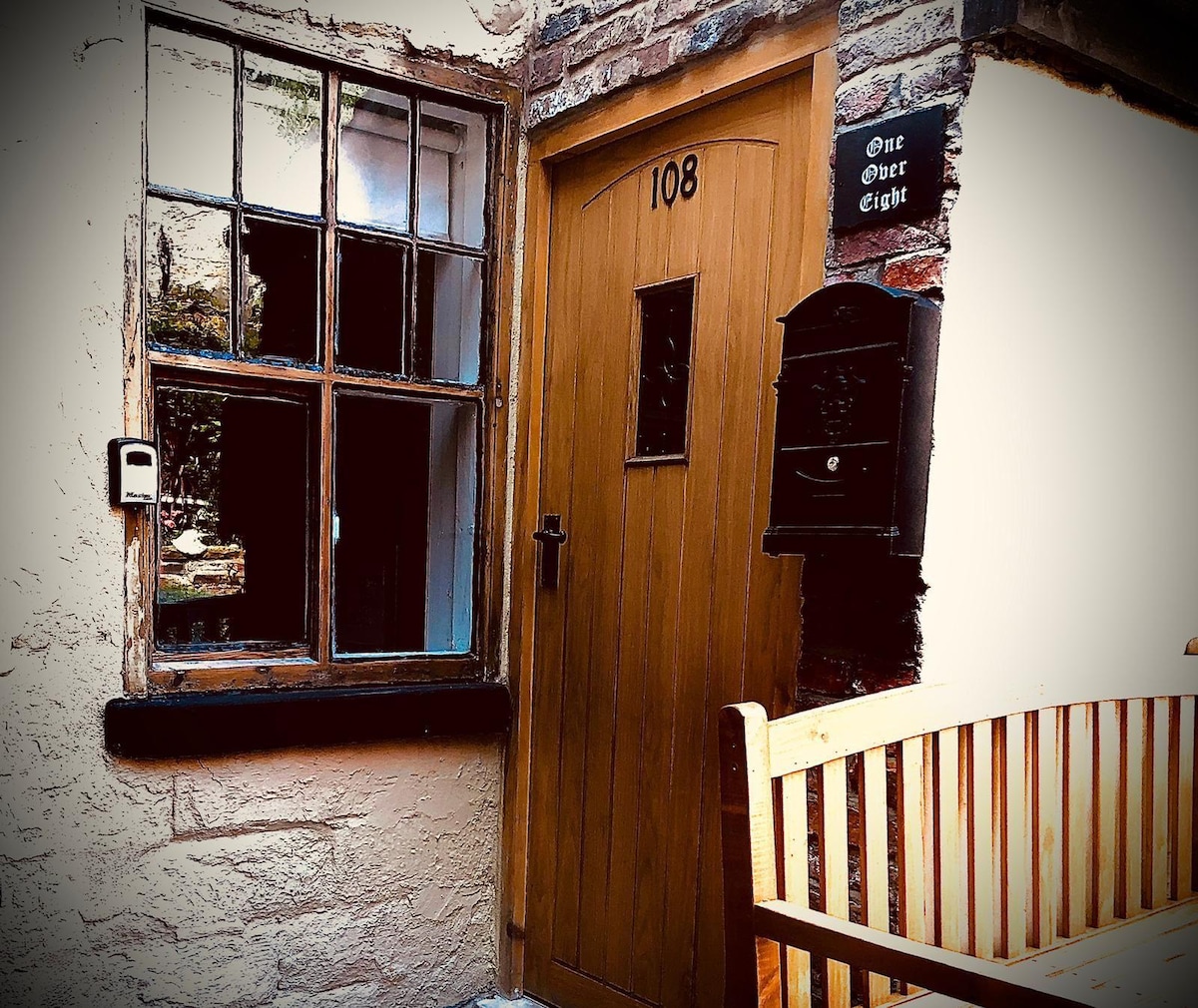 The entrance features a wooden door with a rectangular window, labeled 'One Over Eight.' Beside the door sits a functional mailbox. A wooden bench with a slatted design is positioned in front of the entrance, providing a welcoming spot to relax.