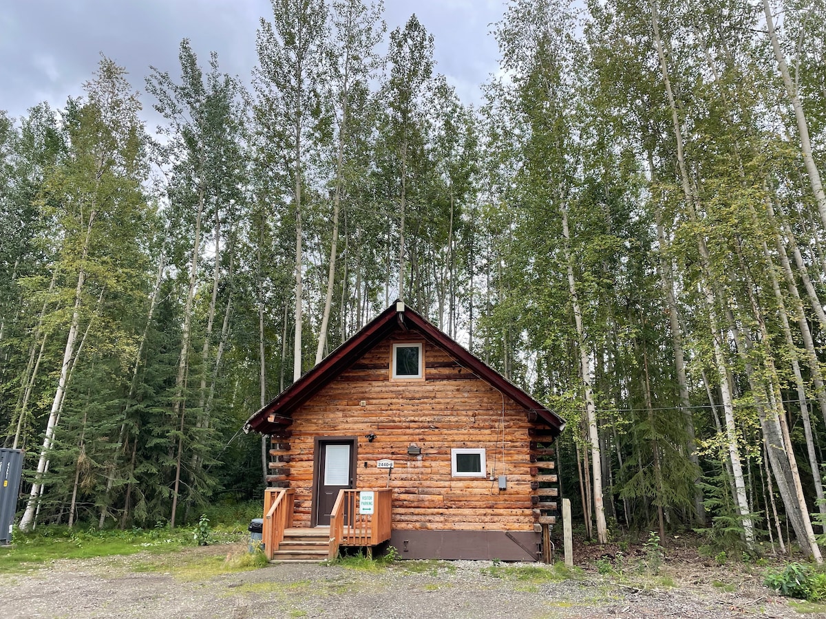 The rustic log cabin is nestled among tall aspen trees, surrounded by greenery. Its wooden exterior features a sloped roof and steps leading to the entrance. Windows are positioned to allow natural light while maintaining privacy. The gravel path offers access to the cabin amidst the tranquil woodland.