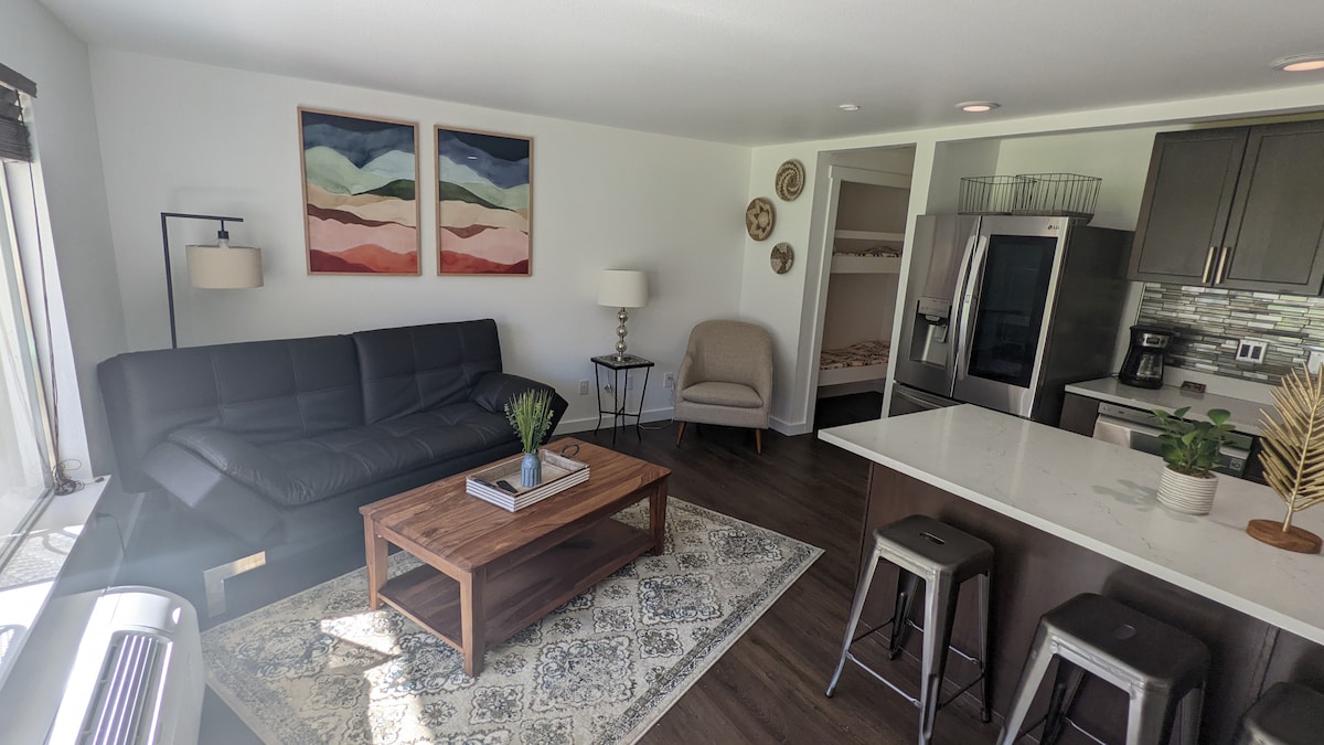 The living area displays a sleek black sofa paired with a light armchair, positioned next to a wooden coffee table. Colorful wall art adorns the white walls. The kitchen features modern appliances and a countertop with bar stools, creating a welcoming space for relaxation.