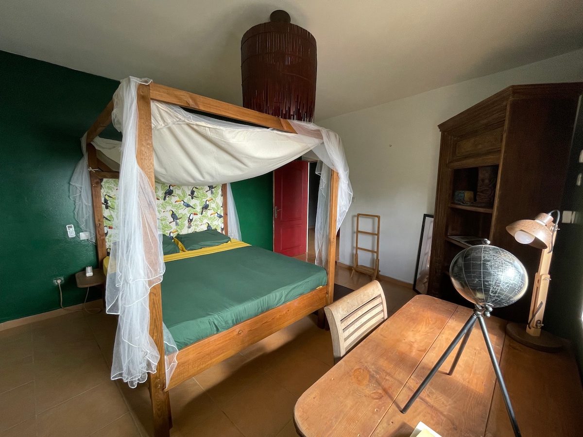 A spacious room features a four-poster bed with a green mattress and soft white drapes. A wooden desk and chair are present, along with a vintage cabinet against a dark green wall. Natural light enters through a nearby window, enhancing the calming atmosphere.