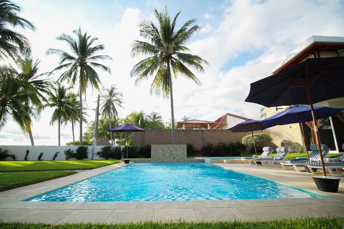 A refreshing pool is surrounded by well-maintained grass and tropical palm trees. Spacious lounge chairs are situated around the pool, providing ample space for relaxation. Umbrellas offer shade for guests enjoying the outdoor setting, creating a serene atmosphere for leisure.