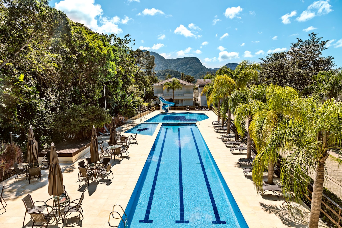 A large outdoor pool is bordered by lush greenery and palm trees. Multiple lounge chairs and umbrellas are positioned along the deck, while a separate area features a smaller pool. The clear blue water reflects the sky, enhancing the inviting atmosphere.