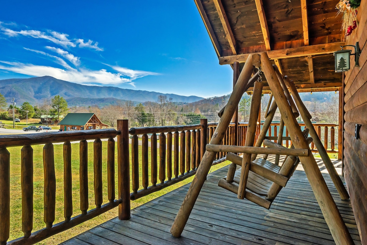 A spacious wooden deck features a large log swing chair, inviting relaxation while overlooking expansive views of the Smoky Mountains. The bright blue sky and distant greenery create a serene backdrop for outdoor enjoyment.