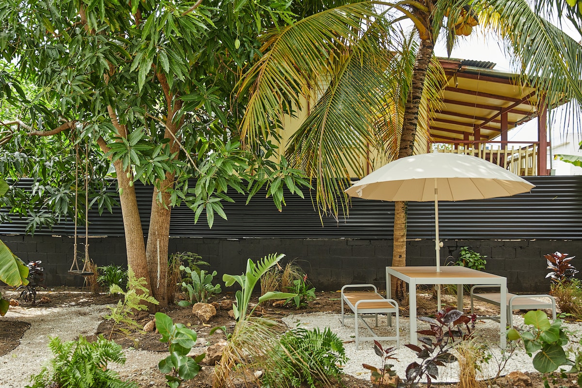 A private garden is surrounded by lush greenery, featuring a dining area with an umbrella and seating. A lightweight table is accompanied by two lounge chairs, while a hammock and a swing are nestled among the trees, providing a serene outdoor retreat.