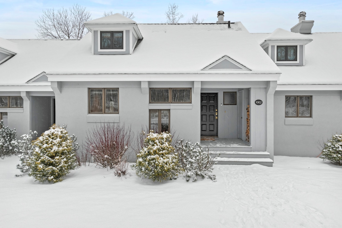A welcoming exterior of a mountain retreat is showcased under a blanket of snow. The entrance features a solid front door, flanked by two symmetrical windows. Snow-covered shrubs and a pathway leading to the door emphasize the serene winter landscape.