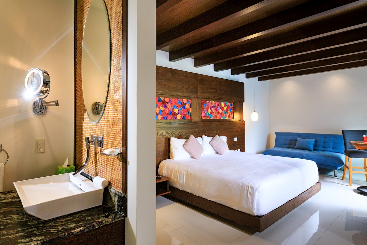 A spacious suite features a comfortable bed with decorative pillows, complemented by wooden accents. The bathroom area includes a stylish sink and a large mirror. A blue couch and a dining area are visible, enhancing the room's inviting layout.