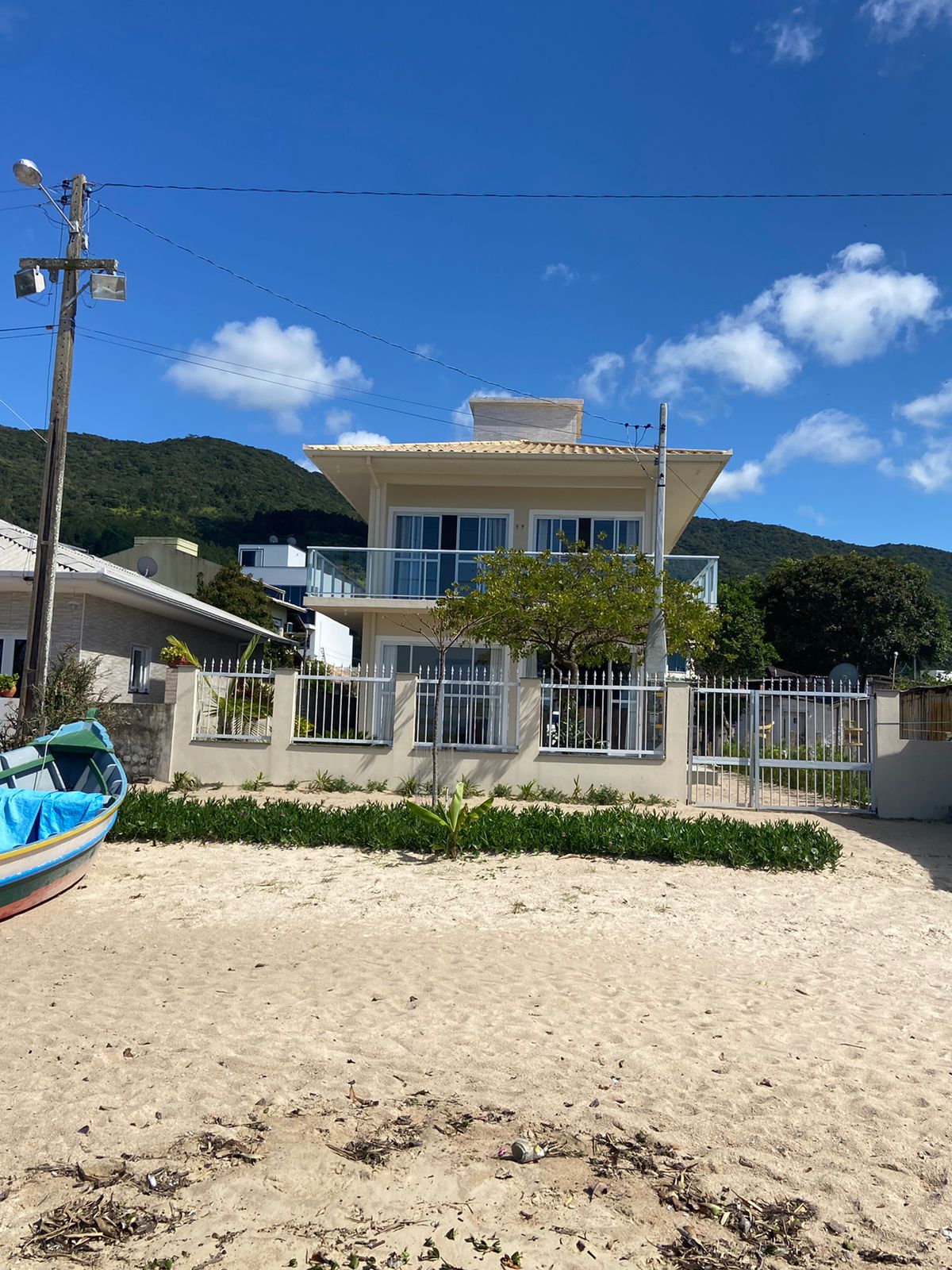 The house is situated on the beach, featuring a modern two-story exterior with a front garden. Large windows provide a view of the surroundings. The sandy area in front is complemented by a small boat, creating a relaxed coastal atmosphere.