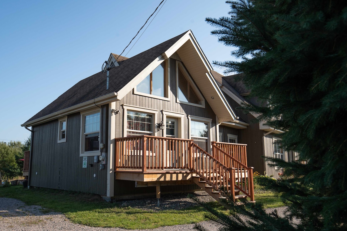 The exterior of a chalet is presented, featuring natural wood siding and a prominent sloped roof. A welcoming front deck with a wooden railing extends from the entrance, surrounded by greenery and gravel pathways. Large windows are positioned to allow ample natural light.