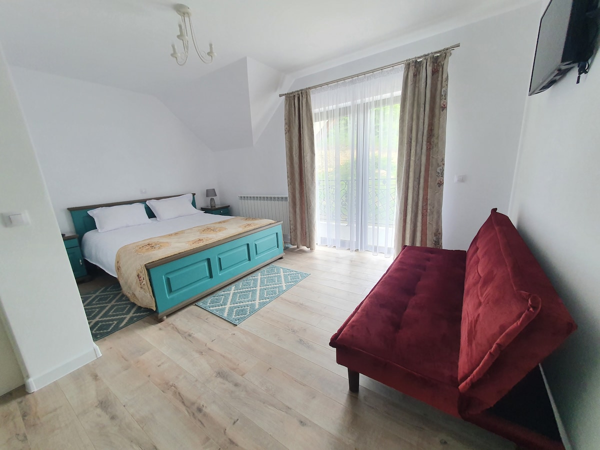 A bright sleeping area features a spacious bed with a teal-colored base, complemented by a cozy red sofa. Soft natural light enters through the large window, adorned with sheer curtains, and a patterned rug adds warmth to the wooden floor.