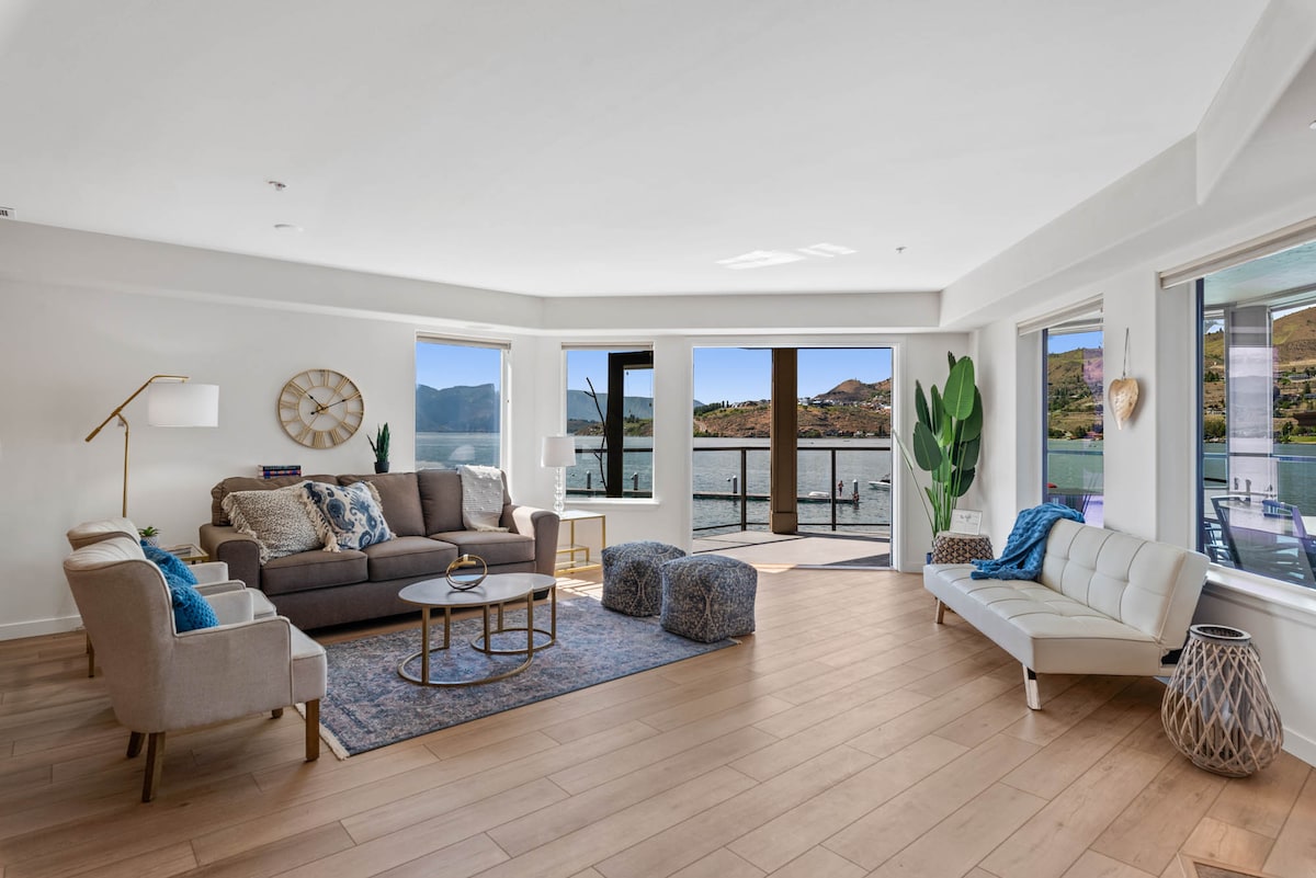 A bright living area features large windows offering lake views and ample natural light. Comfortable seating arrangements include a sofa and two armchairs, complemented by a decorative rug. A coffee table centers the space, and a stylish plant adds a touch of greenery.
