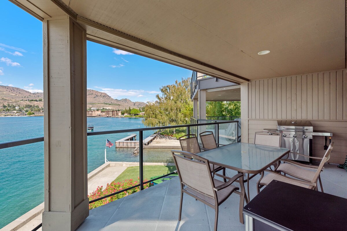 A spacious balcony features a glass table surrounded by several chairs, providing a clear view of the lake and surrounding landscape. A grill is visible nearby, and the scene is framed by moderate greenery and the distant cityscape.
