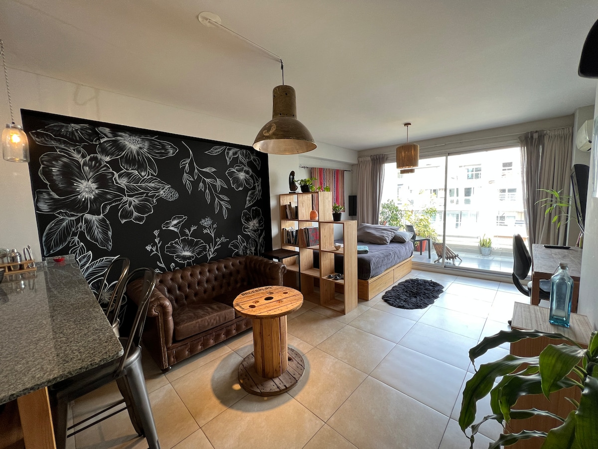 A spacious living area features a large window allowing natural light to fill the room. A brown leather couch is paired with a rustic wooden coffee table. A decorative bookshelf holds books and plants. A comfortable workspace is set up with a chair at a table.