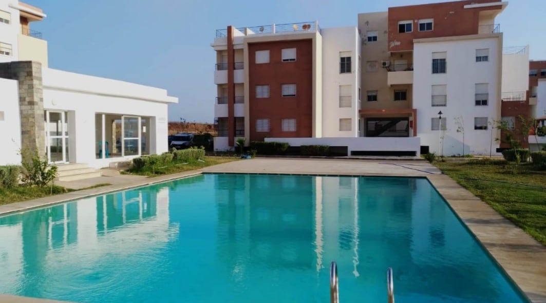 A clear swimming pool is surrounded by landscaped greenery, with modern apartment buildings in the background. The pool area features a well-maintained deck and gentle steps leading into the water. Sunlight reflects off the surface, creating a serene atmosphere.