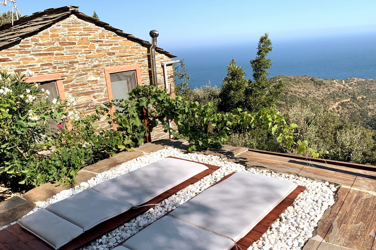 A sun-drenched outdoor area features two lounging mats positioned on a deck surrounded by white pebbles. Lush greenery and blooming flowers frame the space, with a stunning view of the sea and mountains visible in the background.