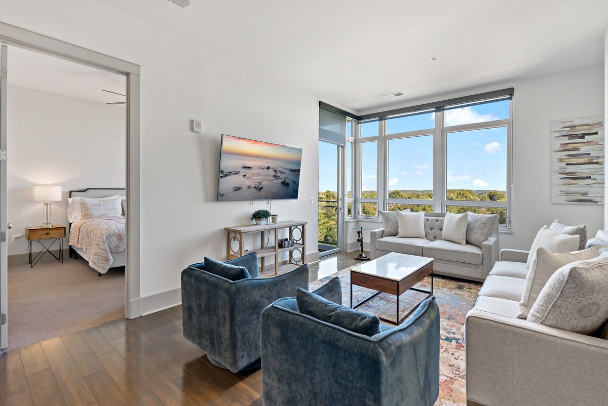 An inviting living space is presented with plush seating arrangements and a coffee table, perfect for relaxation. A smart TV is mounted on the wall, while large windows allow natural light to illuminate the area, offering views of the surrounding landscape.