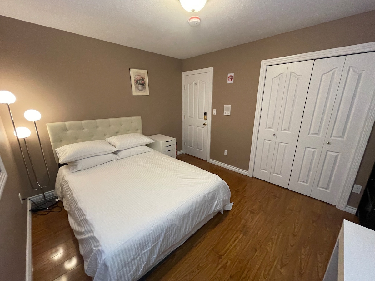 A calm bedroom features a spacious bed with white linens and a light-colored headboard. Two bedside lamps provide soft lighting beside a small nightstand. A large closet with double doors is visible along one wall, and a door leads to the hallway.