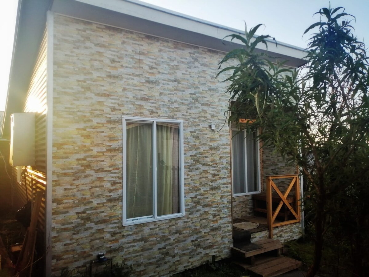 A charming exterior of a small house features a textured stone wall. A wooden staircase leads to the entrance, framed by lush greenery. Large windows allow natural light to enter, while soft curtains are gently drawn, creating a serene atmosphere.