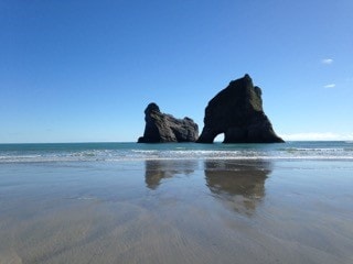 The image captures a scenic beach scene, featuring two large rock formations rising from the water. The clear blue sky reflects off the calm ocean surface, while gentle waves lap at the sandy shore. The symmetry of the rock formations adds to the tranquil atmosphere.