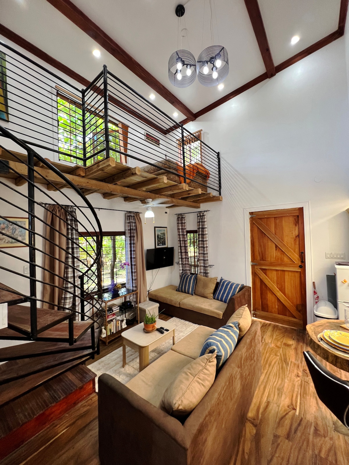 An open living space features a spiral staircase leading to a loft area. Two comfortable sofas face each other, with a coffee table positioned centrally. Large windows allow natural light to fill the room, while soft lighting from stylish fixtures enhances the ambiance.