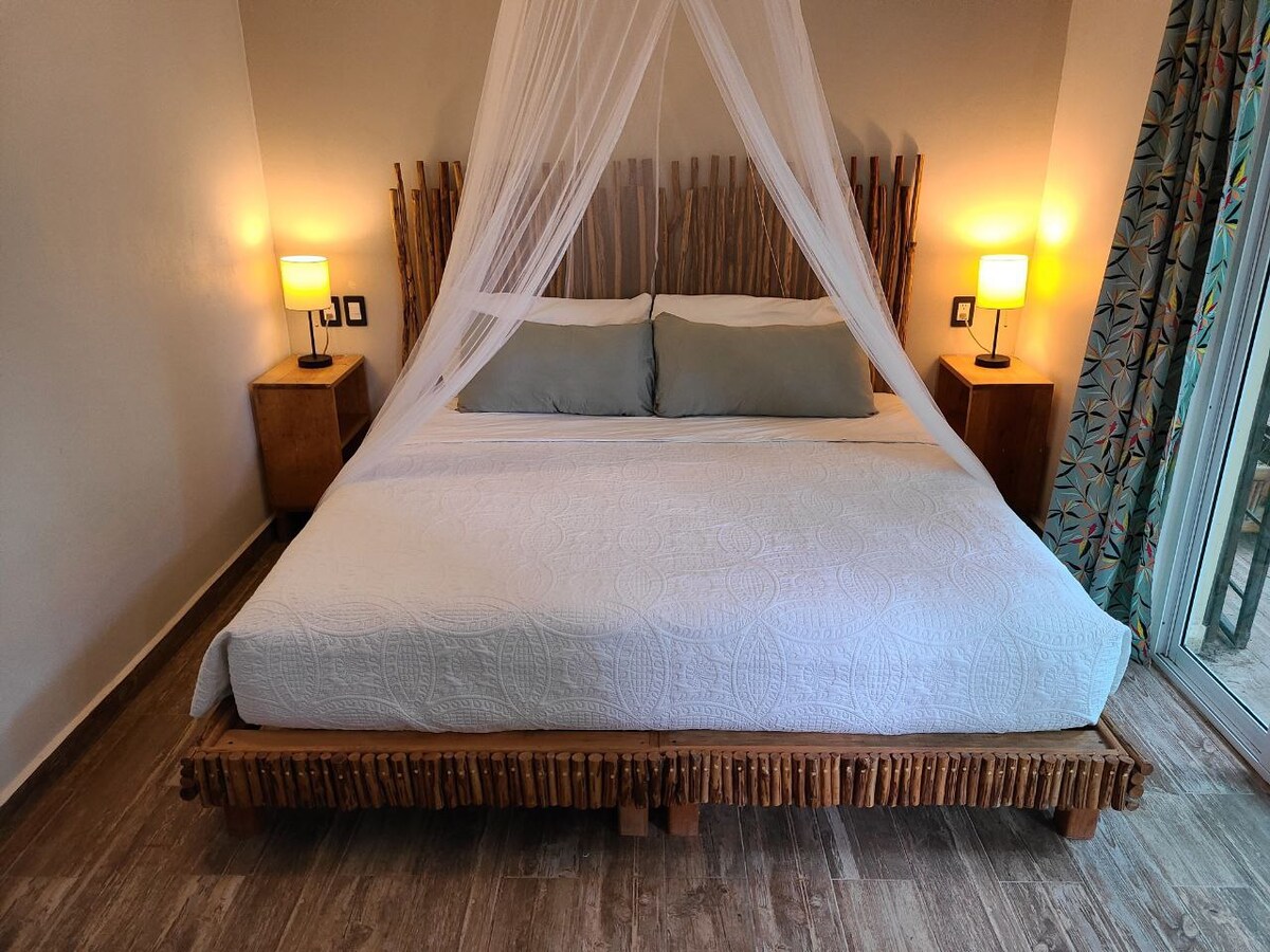 A spacious bed is framed by natural wood accents, featuring light-colored bedding and two decorative pillows. Soft bedside lamps emit a warm glow on either side, while sheer drapery creates an airy feel. A sliding glass door opens to the outdoor area, adding natural light.