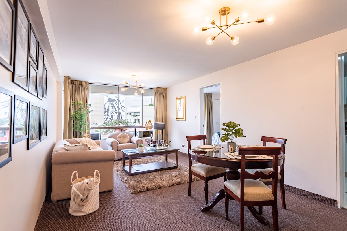 A spacious living area is highlighted by modern light fixtures and large windows providing natural light. A comfortable sofa and accent chairs surround a glass coffee table, while a dining table is positioned nearby. The decor includes framed artwork and indoor plants, creating a calming atmosphere.
