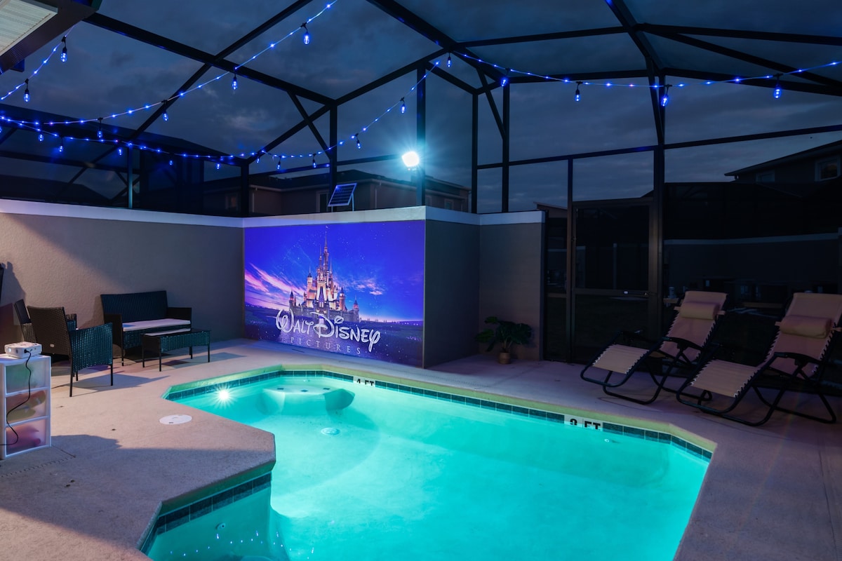 A private pool area is illuminated by string lights, creating a festive ambiance. A large movie projector screen displays a Disney-themed image on a wall. Comfortable seating and sun loungers are arranged nearby, inviting relaxation and enjoyment.