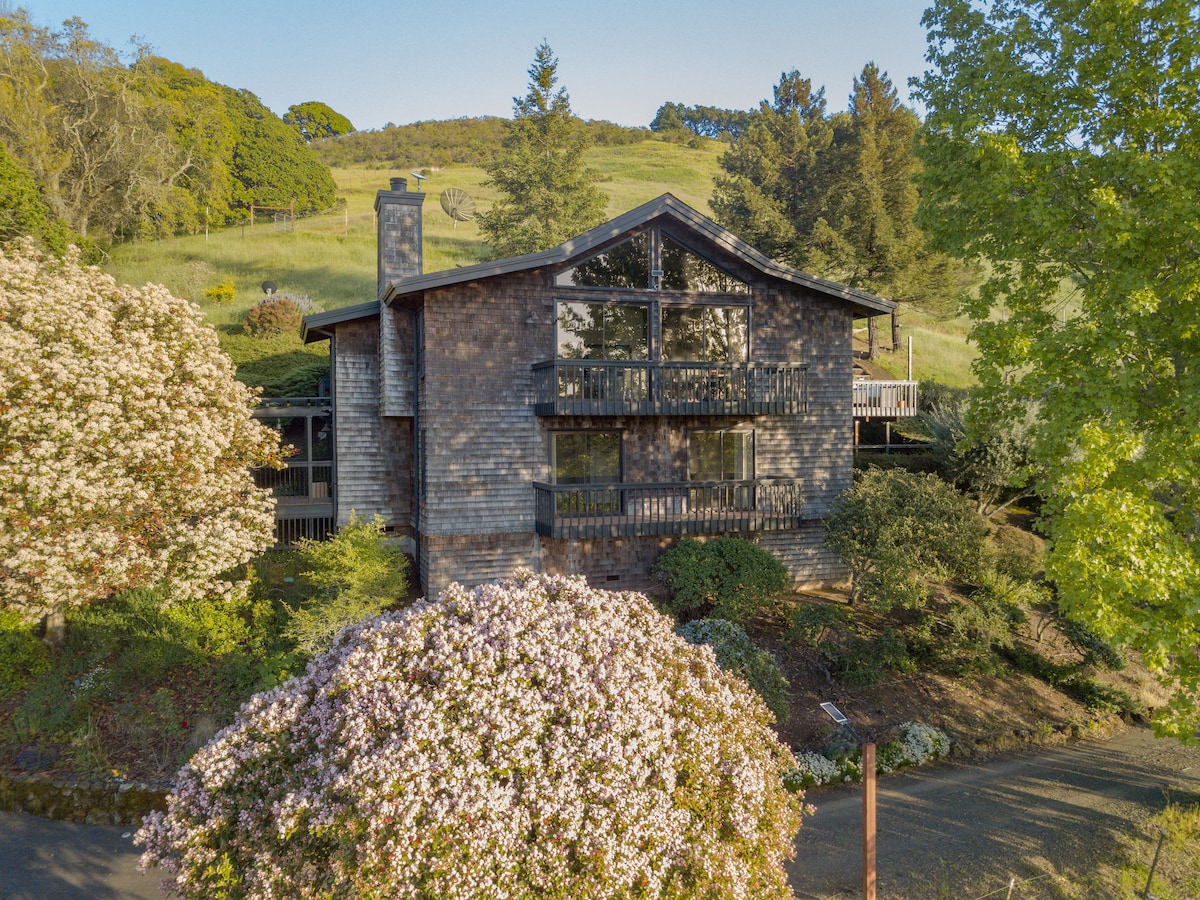A two-story modern farmhouse is surrounded by blossoming trees and lush greenery, highlighting its large windows that invite natural light. The home is perched on a hillside, offering scenic views of the landscape and gardens.