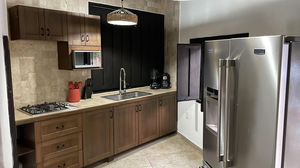 The kitchen features wood cabinetry complemented by a natural stone accent wall. A stainless steel refrigerator and a built-in oven are visible. A countertop with a double sink and a gas stove provide ample preparation space. Small appliances are neatly arranged on the counter.