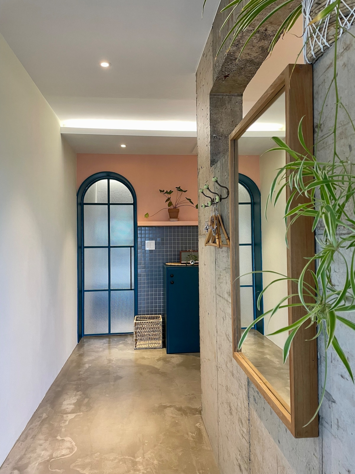 A bright corridor showcases a blend of soft pink and blue tones, with a large mirror framed in wood. A geometric glass door adds character, while green plants provide a touch of nature. A minimalist kitchenette is visible, enhancing the overall spacious feel.