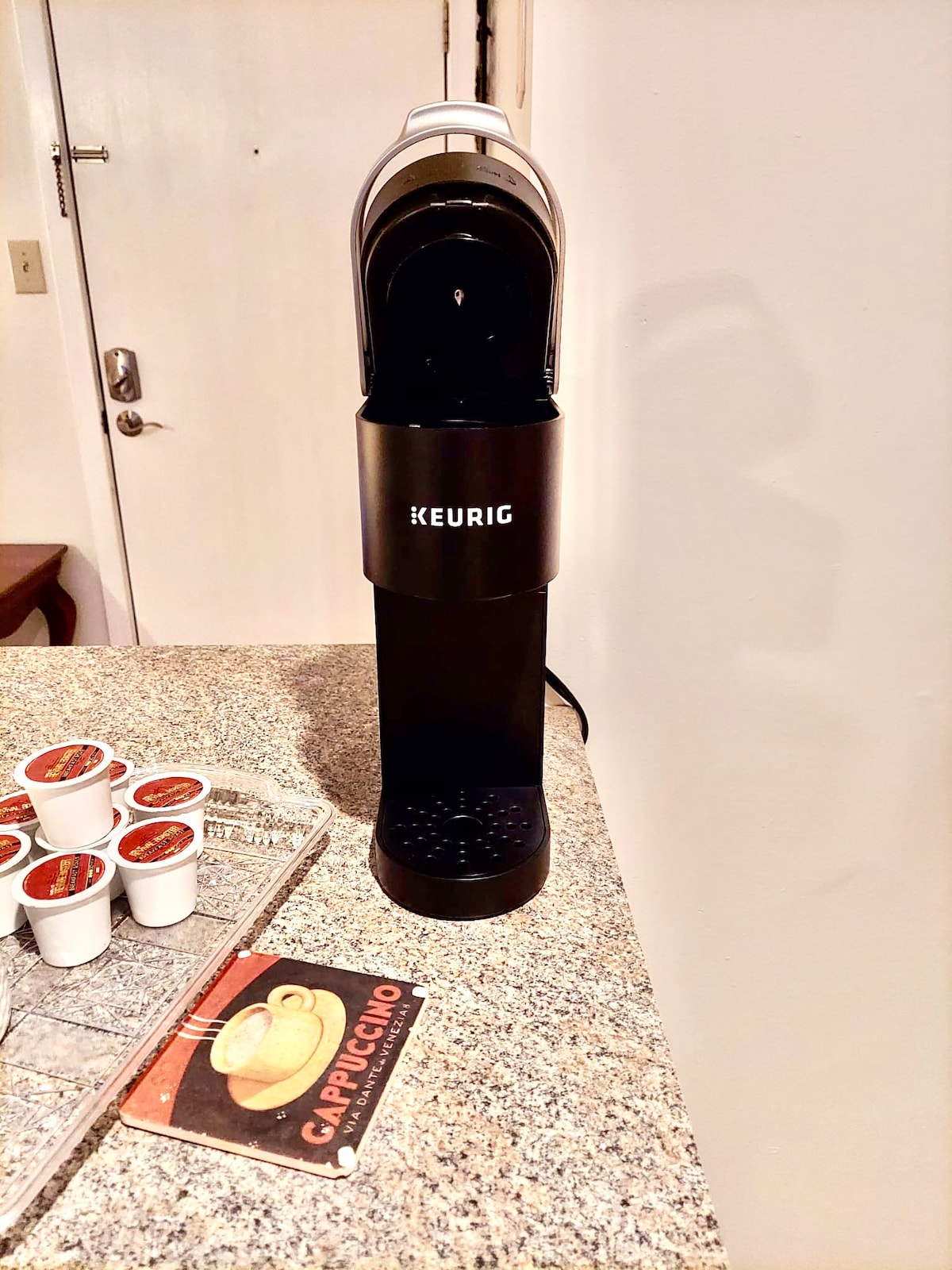 A black Keurig coffee maker is positioned on a textured granite countertop, accompanied by a carousel of coffee pods in various flavors. An informative cappuccino card is placed beside it, enhancing the kitchenette's coffee-making area.