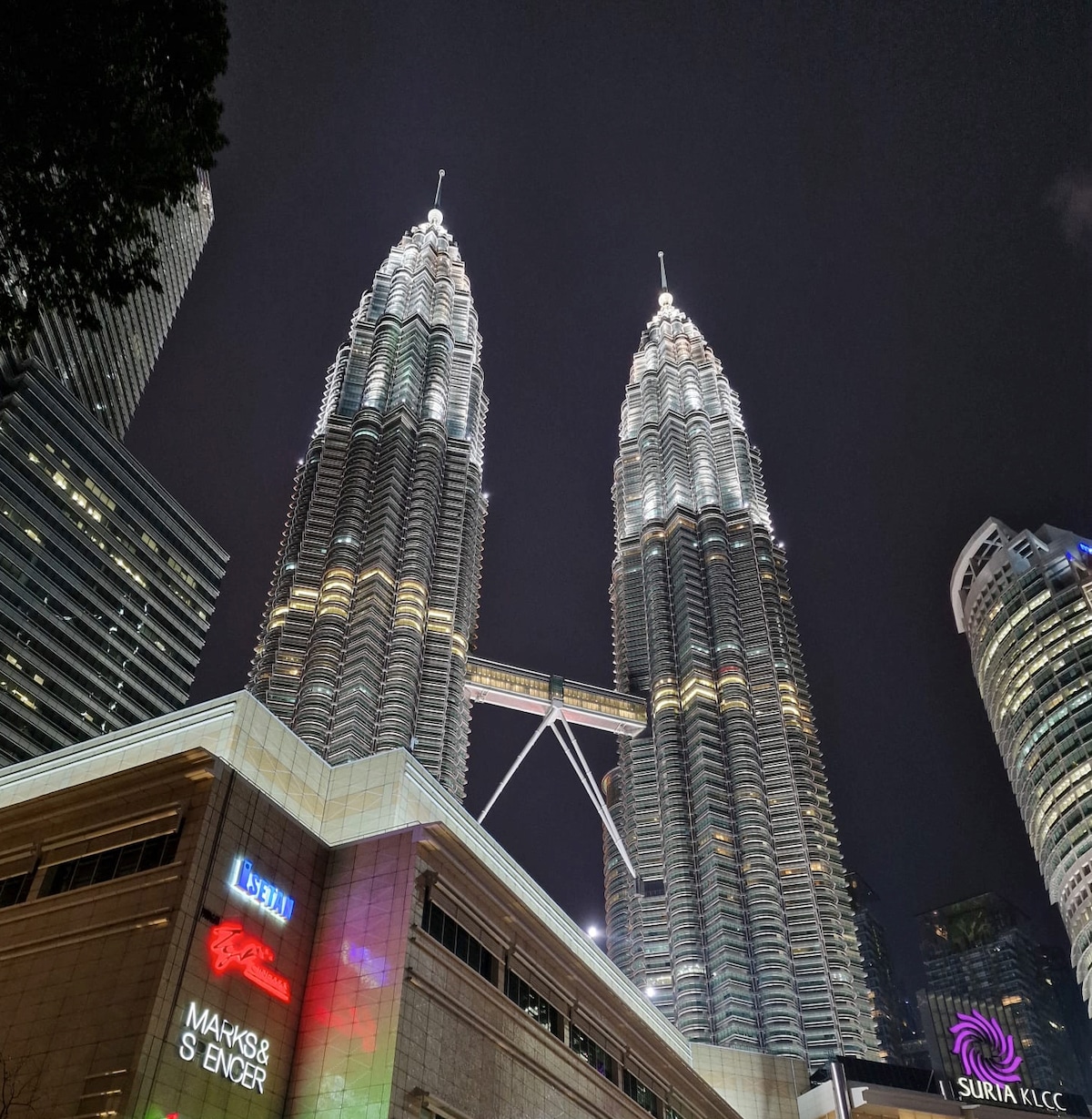 The iconic KLCC Twin Towers are illuminated against a night sky, showcasing their striking architectural features. The surrounding area displays modern buildings, highlighting the vibrant urban landscape. Subtle reflections can be seen on the surfaces of nearby structures, enhancing the scene's lively atmosphere.