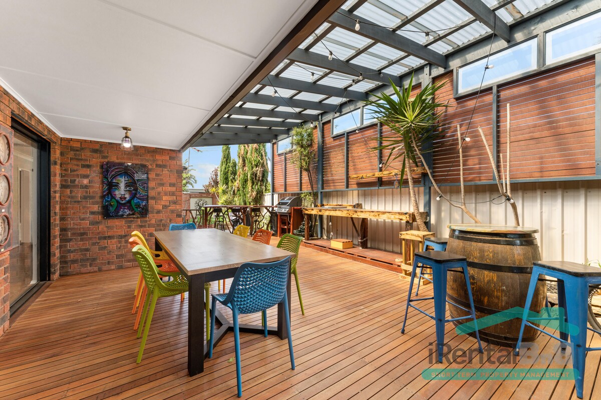 An expansive outdoor deck features a long wooden table surrounded by colorful chairs. A large barrel and barstools provide additional seating. The space is partially covered with a transparent roof, showcasing greenery and a relaxed atmosphere, ideal for gatherings.