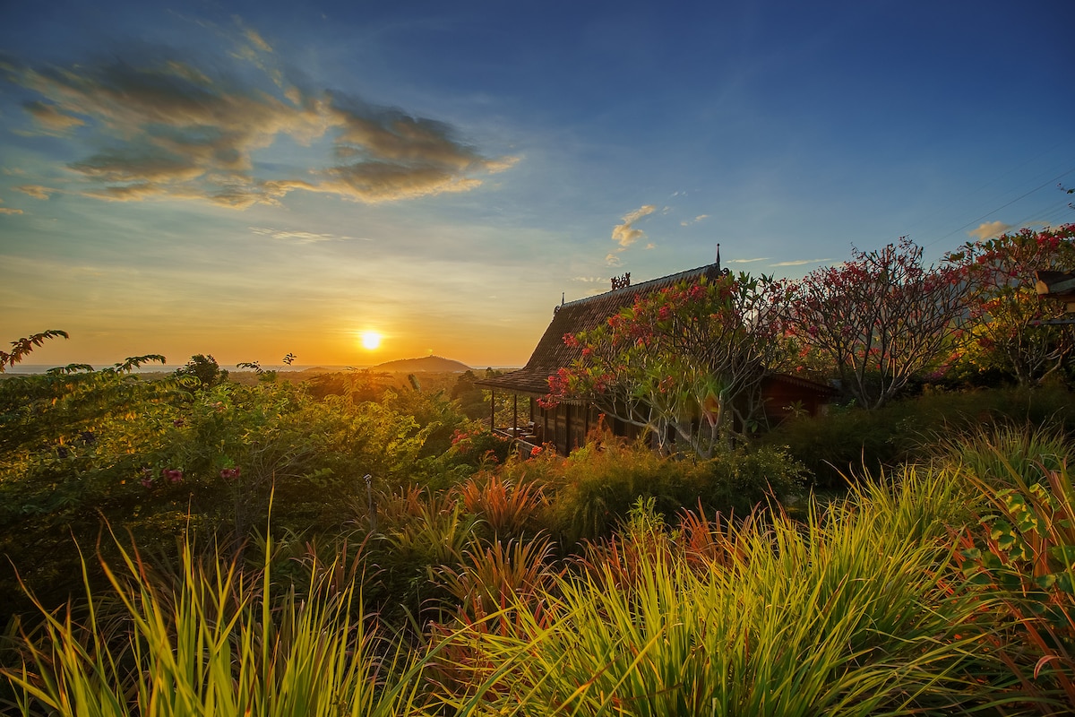 A vibrant sunset paints the sky with warm hues as it sets behind a traditional Javanese house. Lush greenery and colorful flowers frame the scene, highlighting the natural beauty of the retreat's surrounding landscape.
