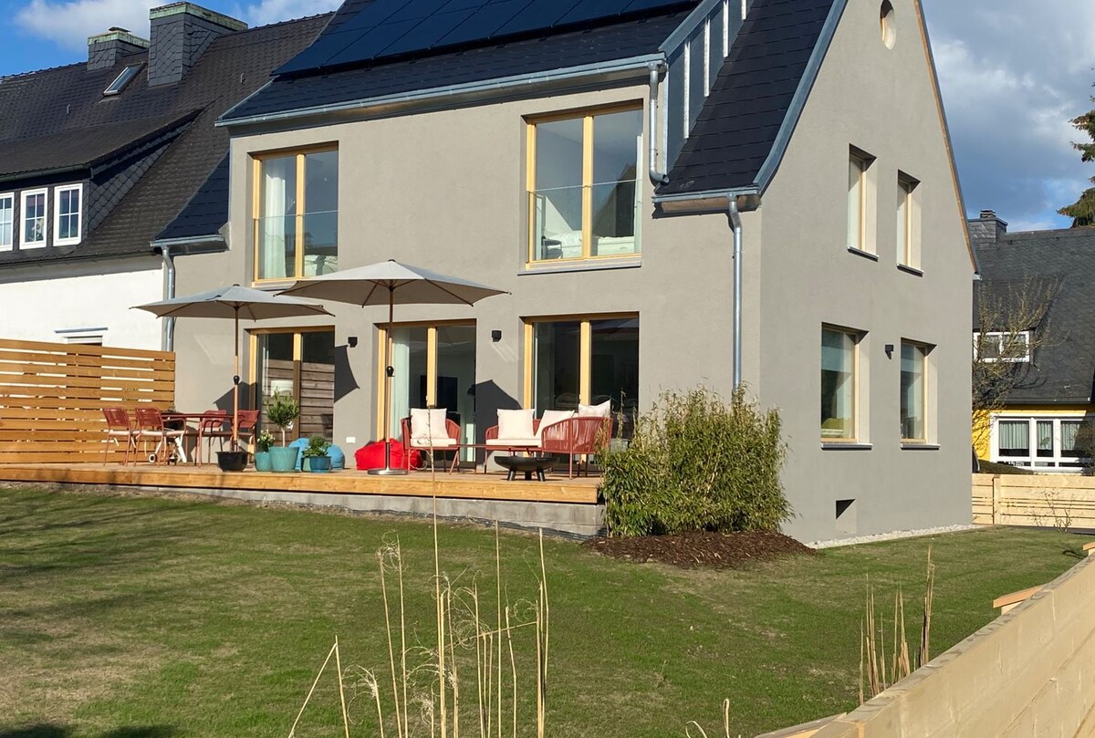 A modern house with large windows features a natural wood deck. Two umbrellas provide shade for outdoor seating arrangements, including several chairs and small tables. The surrounding lawn is well-maintained, and nearby wooden fencing adds a sense of privacy.