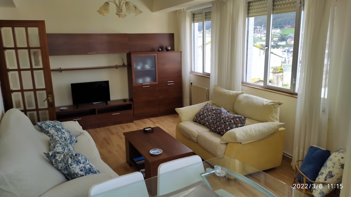 A bright living area is visible, featuring two comfortable sofas, a wooden coffee table, and a television unit with a modern flat-screen TV. Large windows allow natural light to fill the space, showcasing views of the surrounding area.